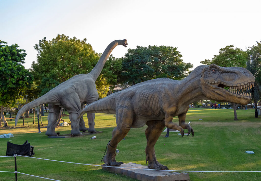 Lebensgroße Dinosauriermodelle in einem Park, darunter ein Tyrannosaurus Rex und ein Brachiosaurus.