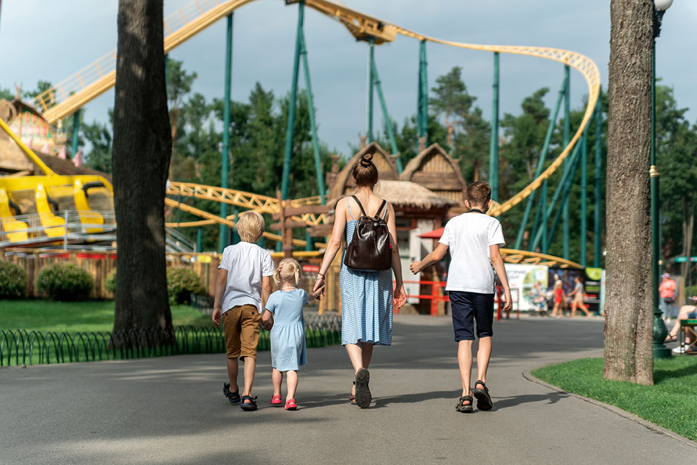 Familie mit drei Kindern spaziert Hand in Hand im Freizeitpark, im Hintergrund Achterbahnen.